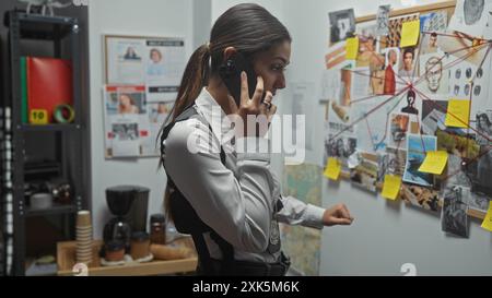 Detective ispanica al telefono in un ufficio investigativo con una scheda delle prove Foto Stock