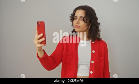 Giovane donna ispanica che scatta un selfie con un telefono rosso, isolata su uno sfondo bianco con giacca rossa e top bianco. Foto Stock