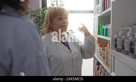 Un'anziana donna bionda caucasica interagisce con un cliente in farmacia, indossando un cappotto bianco e stando vicino a scaffali pieni di vari prodotti Foto Stock