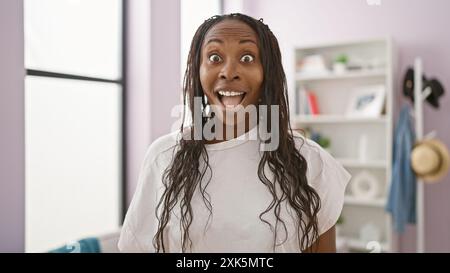 Una donna afroamericana sorpresa con capelli ricci al chiuso, in un soggiorno con uno scaffale sullo sfondo. Foto Stock