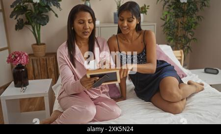 Madre e figlia sedute insieme su un letto in un'accogliente camera da letto, condividendo un momento di amore guardando una cornice fotografica, circondate da piante e casa Foto Stock