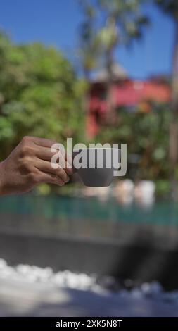 Mano dell'uomo che tiene una tazza di caffè in ceramica all'aperto con sfondo tropicale sfocato e luce solare. Foto Stock