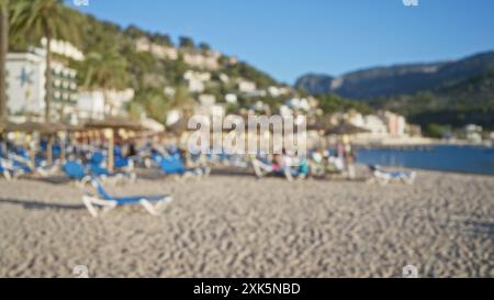 Spiaggia sfocata a maiorca con persone che si godono una giornata di sole su sdraio sotto ombrelloni di paglia, montagne ed edifici sullo sfondo. Foto Stock