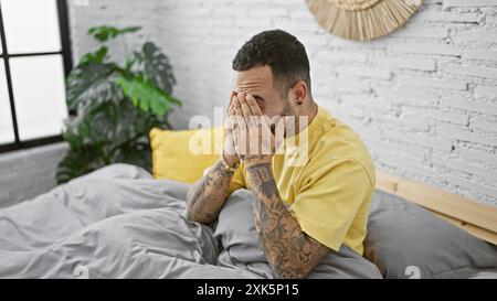 Un angosciato ispanico con tatuaggi copre il viso con le mani, seduto in una camera da letto in stile mattone bianco. Foto Stock