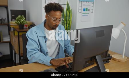 Bel giovane afroamericano con la barba che lavora diligentemente in un computer in un ufficio moderno con piante e grafici sullo sfondo. Foto Stock