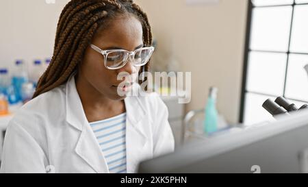 Donna afroamericana concentrata con trecce che lavora in un laboratorio scientifico al chiuso Foto Stock