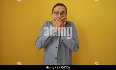 Uomo di mezza età isolato su uno sfondo giallo copre la bocca con entrambe le mani in sorpresa o in shock, indossando occhiali e una camicia grigia. Foto Stock