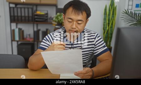 Bel giovane cinese che lavora in un ufficio legge un documento con attenzione tenendo gli occhiali, uomo adulto asiatico concentrato in un posto di lavoro al chiuso W Foto Stock