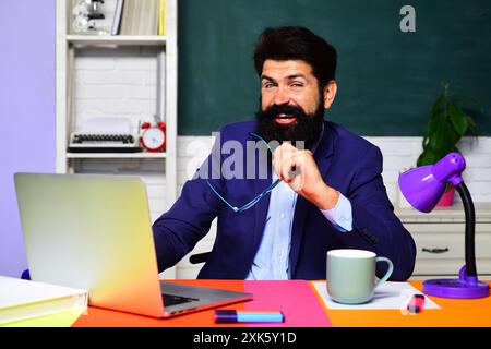 Insegnante di scuola sorridente o tutor universitario che si siede alla scrivania in classe. Torna a scuola. L'insegnante universitario lavora con un computer portatile. Studente di sesso maschile che si sta preparando Foto Stock