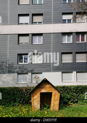 Una piccola casa da gioco in legno si trova in una zona di erba e fiori selvatici di fronte ad un moderno edificio di appartamenti. La sala giochi ha un tetto inclinato e un canto Foto Stock