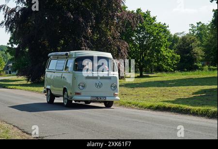 Camper Volkswagen classico che guida attraverso cottage del villaggio e alberi sullo sfondo. Foto Stock