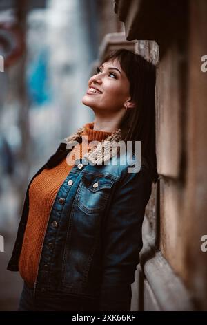Una giovane donna che indossa una giacca in denim e un maglione arancione si appoggia contro una parete di mattoni, sorridendo e guardando verso l'alto. Foto Stock