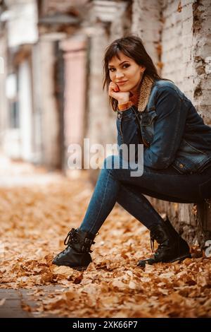Una giovane donna siede contro un muro di mattoni, appoggiata contro la sua mano. Indossa una giacca in denim e un jeans e si siede su un letto di foglie autunnali cadute. Foto Stock
