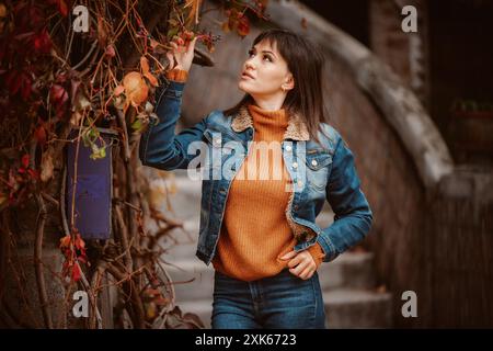 Una donna con giacca in denim e maglione arancione si erge accanto a un albero con viti. Foto Stock