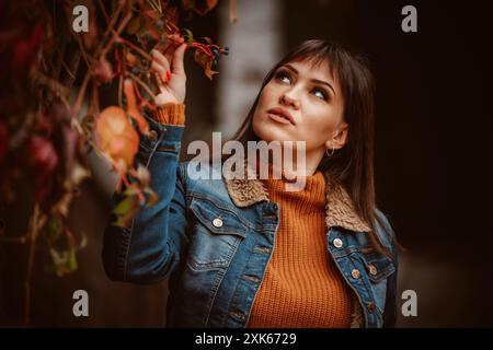 Una donna che indossa una giacca in denim e un maglione arancione guarda le foglie autunnali. Foto Stock
