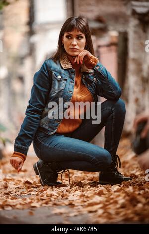 Una giovane donna che indossa una giacca in denim e un jeans si siede su un letto di foglie cadute. La sua mano poggia sul mento mentre guarda di lato. Foto Stock