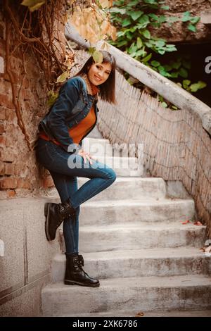 Una giovane donna si appoggia a una parete di mattoni, indossando jeans blu e una giacca in denim mentre guarda la fotocamera. È in piedi su una serie di ste in cemento Foto Stock