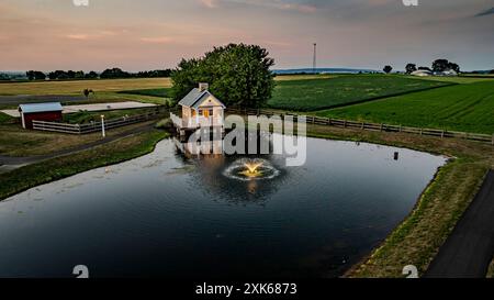 Affascinante scenario rurale al tramonto caratterizzato da Una piccola casa in pietra con Un Portico bianco che si riflette in Uno stagno, circondato da campi verdi, pascoli recintati e Un cielo serale delicatamente illuminato, creando Un ambiente tranquillo. Foto Stock