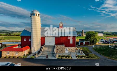 Ronks, Pennsylvania, Stati Uniti, 11 luglio 2024 - grande fienile rosso con tetto d'argento e alto Silo bianco, circondato da giardini ben curati, veicoli e lussureggianti campi verdi sotto Un cielo azzurro con nuvole di Wispy. Foto Stock