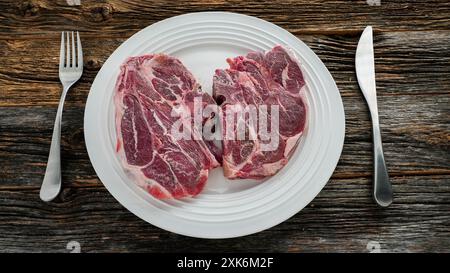 Due costolette di agnello crudo su un piatto bianco con forchetta e coltello su un tavolo di legno Foto Stock