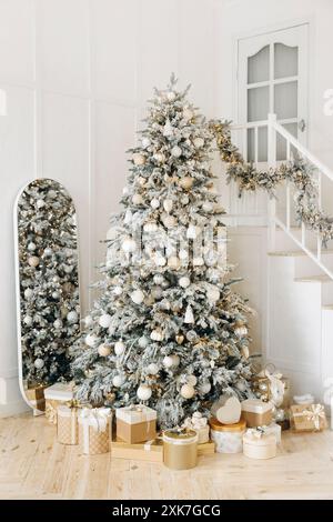 Elegante allestimento natalizio con un albero decorato e una varietà di regali in oro e bianco sotto una scala. La ghirlanda sulle scale e lo specchio aggiungono Foto Stock