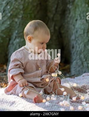 Ritratto di un affascinante bambino di un anno in un abito beige, seduto vicino a un grande albero. Il bambino tiene le lanterne tra le mani e le esamina attentamente Foto Stock