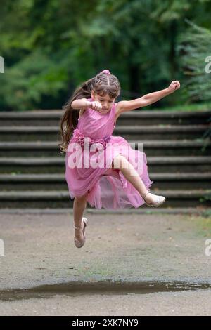 Una bambina con un bellissimo abito rosa dai capelli lunghi corre rapidamente e salta sopra una marionetta. Mi sono concentrato e mi sono morso la lingua. Giochi all'aperto. Sfocato Foto Stock