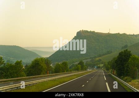 Un'autostrada lunga e dritta si snoda attraverso dolci colline verdi, con una catena montuosa nebbiosa in lontananza. Il sole che tramonta proietta un caldo bagliore sulla terra Foto Stock