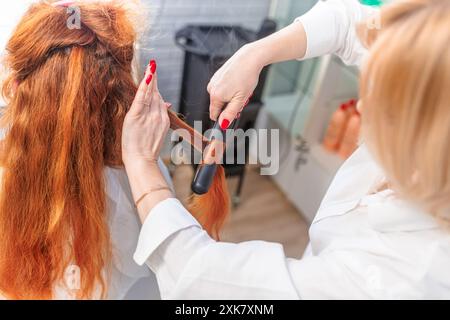 Uno stilista utilizza un ferro da stiro per stirare i lunghi capelli rossi di una donna in un salone. Foto Stock