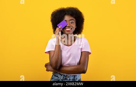 Concetto di acquisto, denaro e pagamento. Gioiosa donna afroamericana che copre l'occhio con carta di credito, sfondo giallo Foto Stock