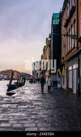 Due persone camminano lungo le strade acciottolate di Venezia, Italia, con canali e architettura storica sullo sfondo. L'atmosfera è tranquilla Foto Stock