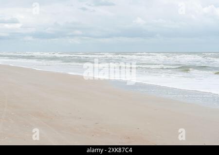 Una spiaggia di sabbia bianca con un mare agitato. Le onde si stanno arrotolando con la schiuma bianca del mare tempestoso. Il litorale vuoto ha un cielo nuvoloso e pallido. Foto Stock