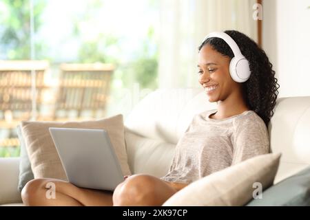 Donna nera felice che usa un laptop e una cuffia sul divano di casa Foto Stock