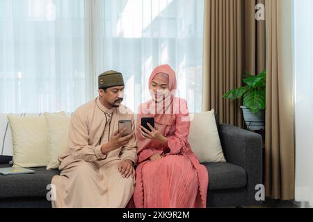 Coppia musulmana asiatica che fa una videochiamata usando un cellulare mentre si siede insieme sul divano durante Eid Mubarak in salotto. Sparato a casa Foto Stock