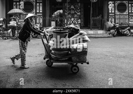 Vecchia vietnamita che spinge un carro con le merci. Hoi An, Vietnam. Una donna che indossa abiti tradizionali vietnamiti spingendo un trolley in metallo. Foto di viaggio i Foto Stock