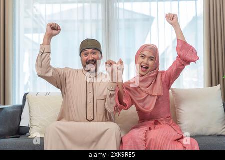 Felice coppia araba in abiti islamici seduti sul divano in soggiorno che mostra gioia con le mani in alto e facce sorridenti, coppia felice che riceve buone notizie. Foto Stock