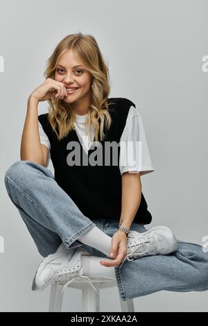 Una giovane donna con i capelli biondi in un abbigliamento elegante si mette in posa mentre si siede su uno sgabello su uno sfondo grigio. Foto Stock