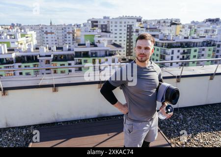 Felice barbuto sportivo e bello uomo che va in palestra o pratica yoga sulla strada della città con un tappetino e una bottiglia d'acqua in mano. cammina Foto Stock