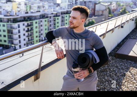 Uomo con barba, sportivo e bello, che ammira la vista della città sul tetto tenendo in mano il tappetino e una bottiglia d'acqua. Foto Stock