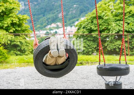 Bambino elementare su un'altalena di pneumatici fatta di catene e pneumatici per auto in un parcheggio. Vista di Bergen, Norvegia Foto Stock