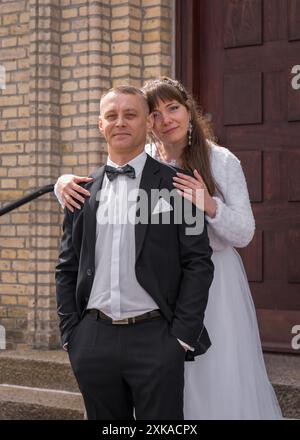 La sposa e lo sposo sono in piedi sui gradini sullo sfondo delle porte marroni. Lo sposo tiene le mani nelle tasche. La sposa sta dietro t Foto Stock