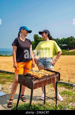 Due donne russe che parlano e cucinano barbecue di maiale sulla spiaggia del mare giapponese in estate Foto Stock