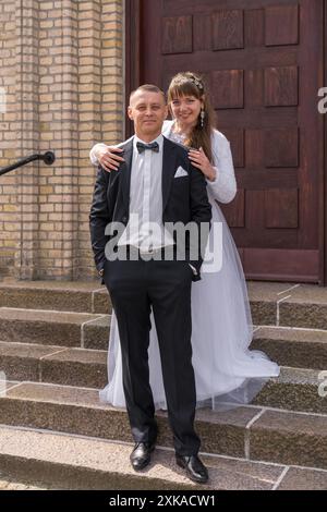 La sposa e lo sposo sono in piedi sui gradini sullo sfondo delle porte marroni. Lo sposo tiene le mani nelle tasche. La sposa sta dietro t Foto Stock