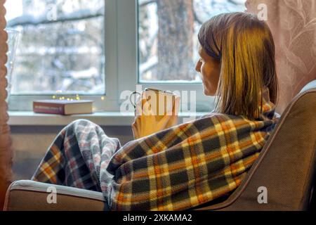 Una donna triste e pensiva siede su una sedia accanto alla finestra con un paesaggio innevato invernale, avvolto in plaid, con tazza ed è annoiato. Solitudine delle vacanze. Foto Stock