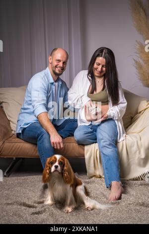 La famiglia sta aspettando un bambino. Un uomo e una donna incinta con una pancia esposta sono seduti e abbracciati sul divano, con un cane seduto accanto a loro Foto Stock