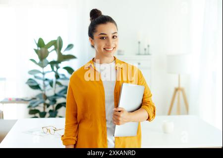 Ritratto di una giovane donna indiana o araba sicura di sé, manager di successo, hr, azienda seo, che tiene in mano un notebook mentre si trova in un ufficio moderno, guarda la fotocamera e sorride Foto Stock