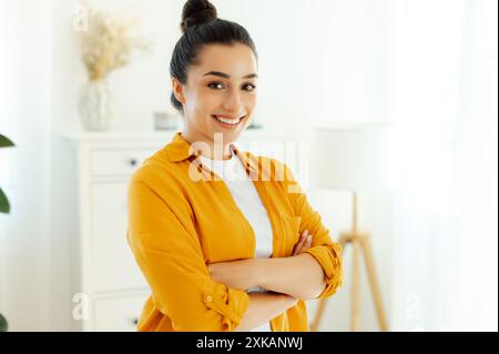 Ritratto di una splendida giovane donna di etnia araba o indiana vestita con camicia arancione, dipendente dell'azienda, direttore creativo si trova all'interno vicino alla scrivania con le braccia incrociate, guarda e sorride alla macchina fotografica Foto Stock