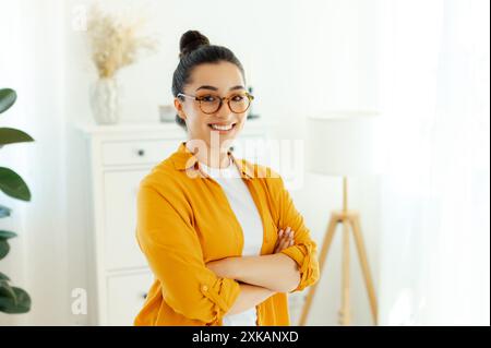 Ritratto di una splendida giovane donna di etnia araba o indiana in camicia arancione con occhiali da vista, dipendente dell'azienda, direttore creativo si trova all'interno vicino alla scrivania, guarda e sorride davanti alla macchina fotografica Foto Stock