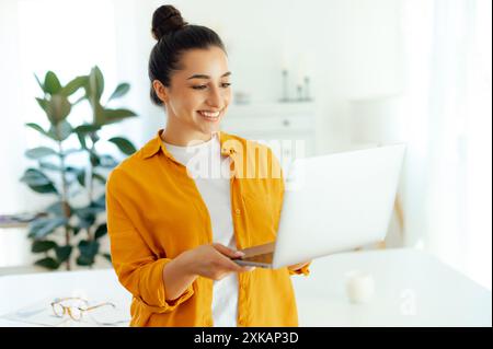 Affascinante donna bruna indiana o araba sicura di sé, manager, copywriter creativo, hr, che tiene in mano un notebook aperto e guarda uno schermo mentre si trova in un ufficio moderno, sorridendo Foto Stock