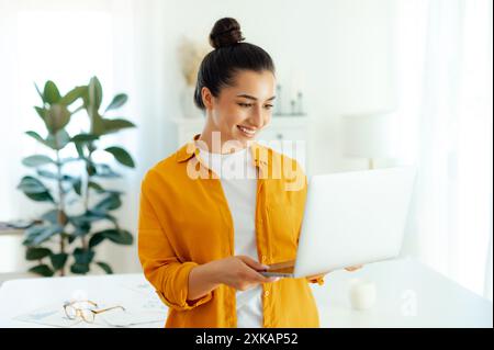 Bella donna bruna indiana o araba di successo, manager, copywriter creativo, hr, che tiene in mano un notebook aperto e guarda uno schermo mentre si trova in un ufficio moderno, sorridendo Foto Stock